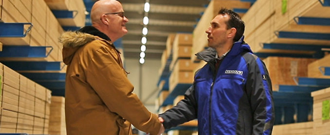 Zwei Personen schütteln sich die Hand. Im Hintergrund Kragarmregale.